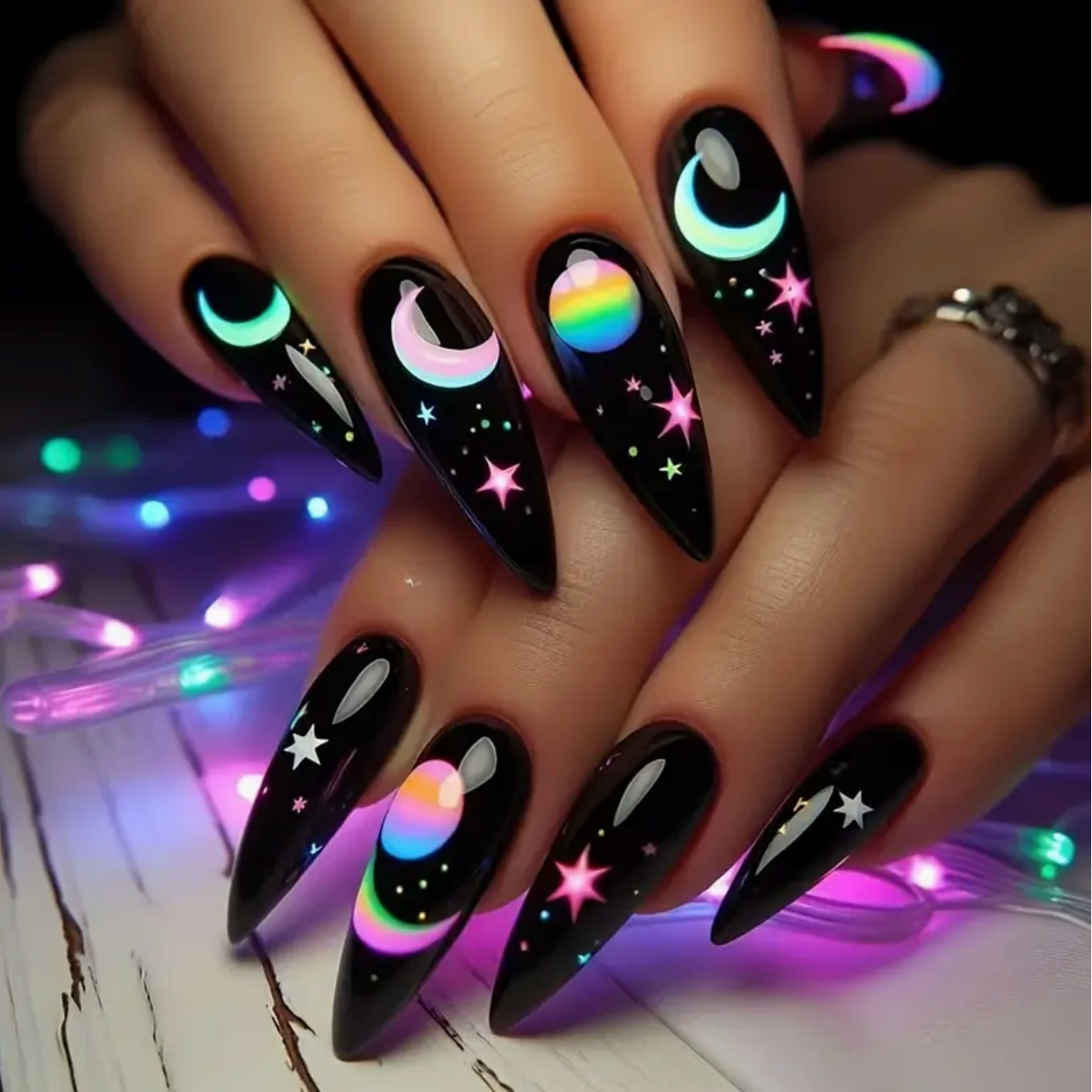 Nails with black polish, colorful designs, and a blurred string of lights in the background.
