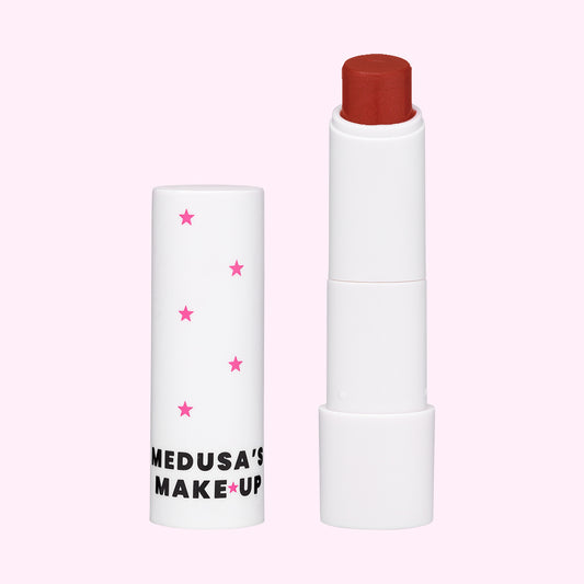 A vegan tinted lip balm by Medusa's Make-up, with the product slightly exposed showing a red color stick, encased in a white tube with pink stars.