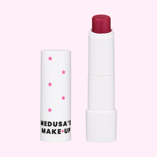 A vegan tinted lip balm by Medusa's Make-up, with the product slightly exposed showing a pink colored balm tip, encased in a white tube with pink stars.