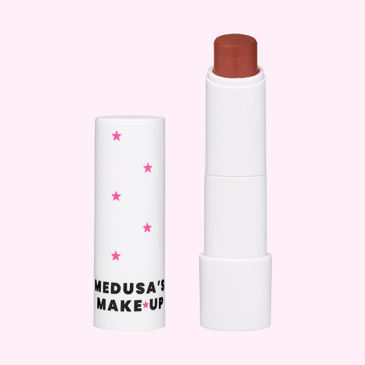 A vegan tinted lip balm by Medusa's Make-up, with the product slightly exposed showing a brown lip balm stick. The container is white with pink stars.