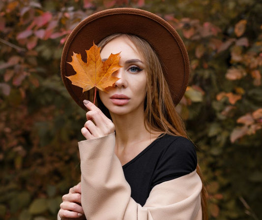 Model wearing a hat posing in fall foliage and holding a maple leaf up to her eye
