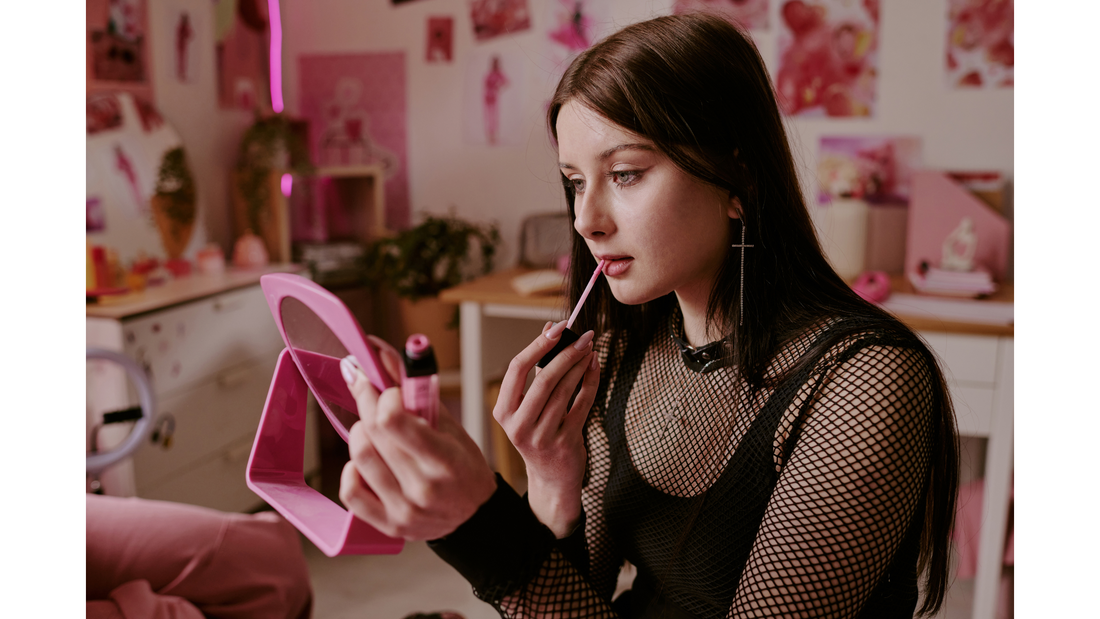 female holding a mirror and applying lipstick, she is wearing a black fishnet material shirt