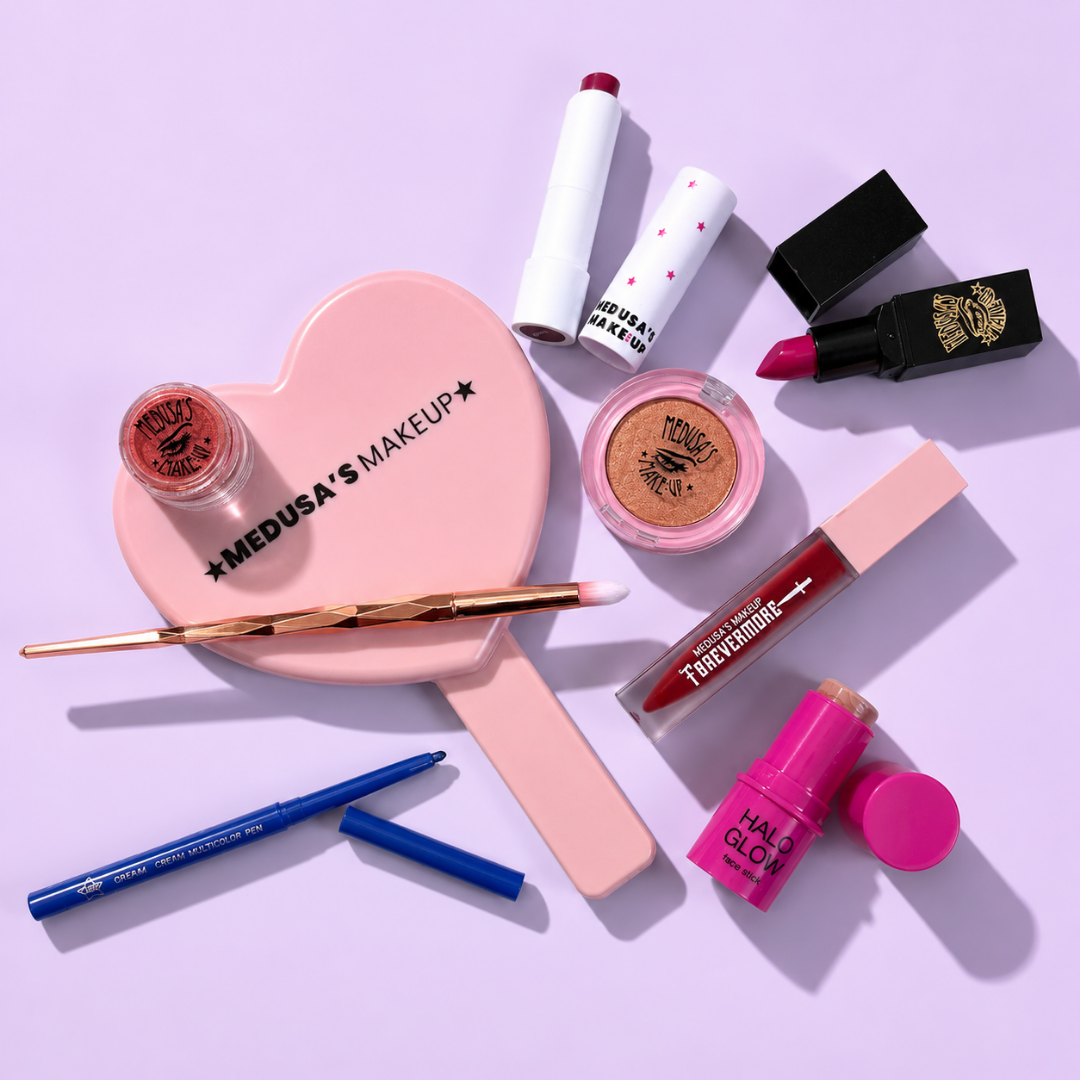 A flat-lay arrangement of beauty products on a light lavender background, including a pink heart-shaped mirror labeled 'MEDUSA'S MAKEUP,' a small eyeshadow pot, a makeup brush, a blue pencil, a pink lip gloss tube, a red lipstick, a gold eyeshadow compact, and other cosmetics.