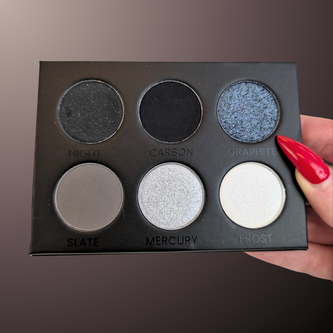 A six-pan eyeshadow palette held by a hand with red manicured nails. The palette is black and contains six shades labeled 'NIGHT', 'CARBON', 'GRAPHITE', 'SLATE', 'MERCURY', and 'FROST' in a matte, shimmer, and shimmer/silver finish.