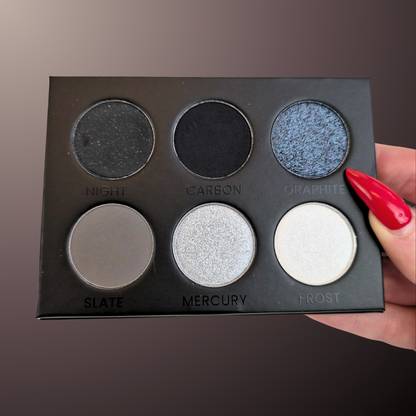 A six-pan eyeshadow palette held by a hand with red manicured nails. The palette is black and contains six shades labeled 'NIGHT', 'CARBON', 'GRAPHITE', 'SLATE', 'MERCURY', and 'FROST' in a matte, shimmer, and shimmer/silver finish.