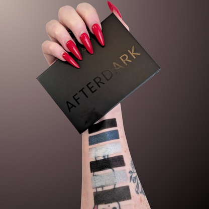 A hand with long red stiletto nails holds a black rectangular makeup palette labeled AFTEDARK. The person's forearm has eyeshadow swatches in black, blue, gray, and white.