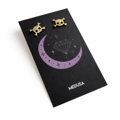 Gold skull-shaped earrings on a black card with 'Medusa' branding.