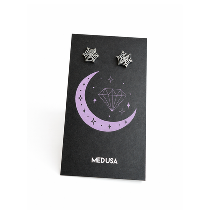 Black card with purple crescent moon design and 'MEDUSA' branding, featuring silver spider web earrings.