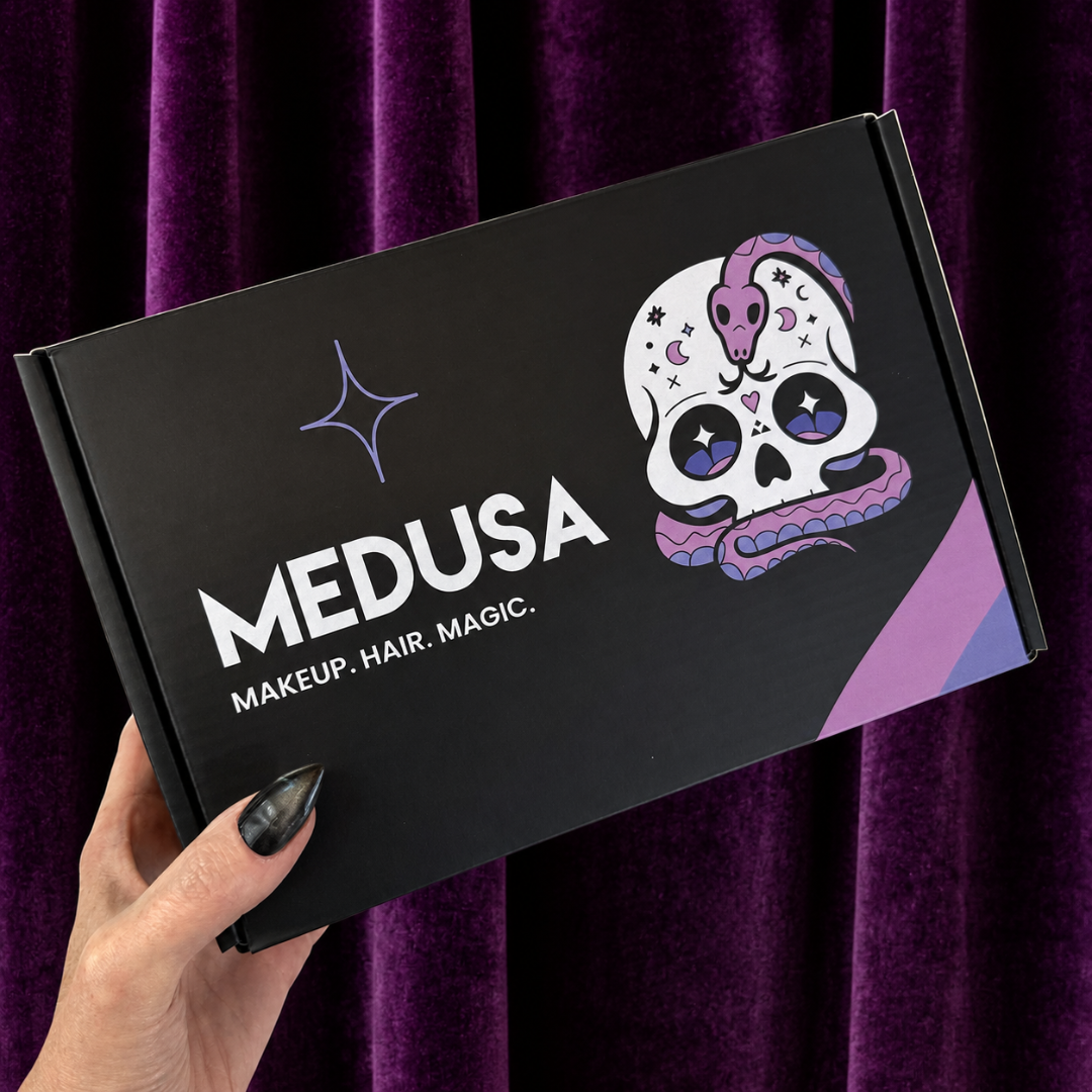 A hand with black nail polish holds a black Medusa makeup, hair, and magic box against a purple velvet curtain background. The box features a white sugar skull illustration with purple and blue details and the brand name 'MEDUSA' in white.