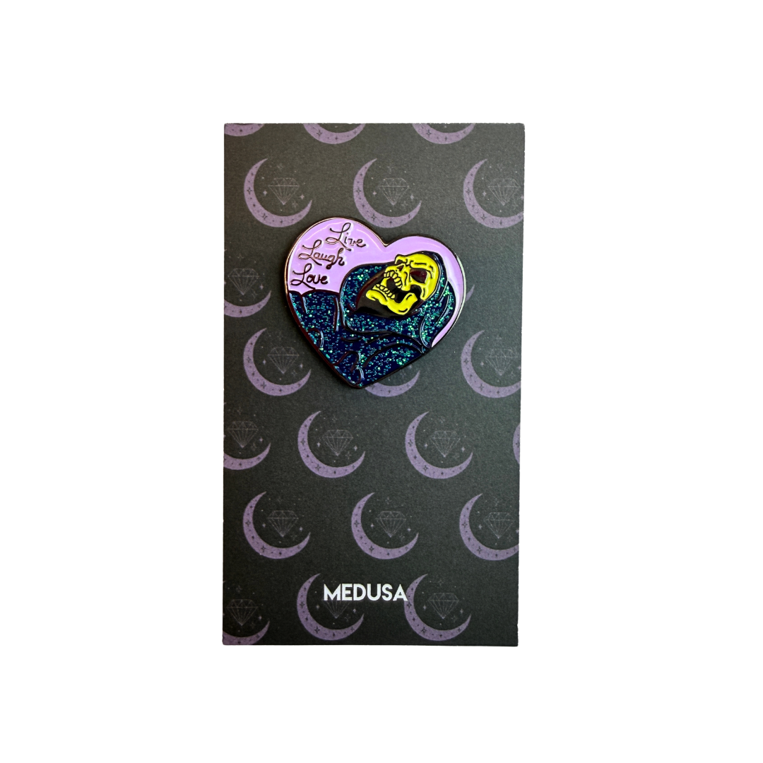 Heart-shaped pin with a yellow face design on a black card with crescent moon patterns, branded 'Medusa'.
