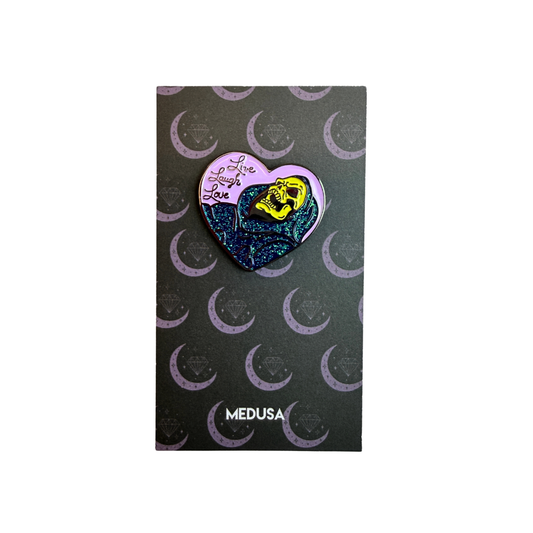 Heart-shaped pin with a yellow face design on a black card with crescent moon patterns, branded 'Medusa'.