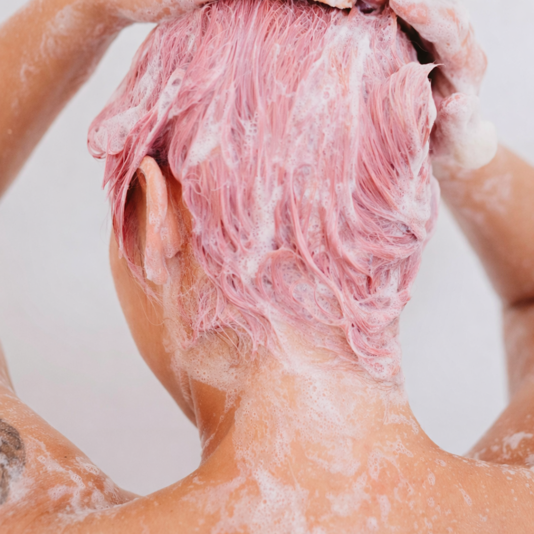 Person with pink hair washing their hair with shampoo against a neutral background
