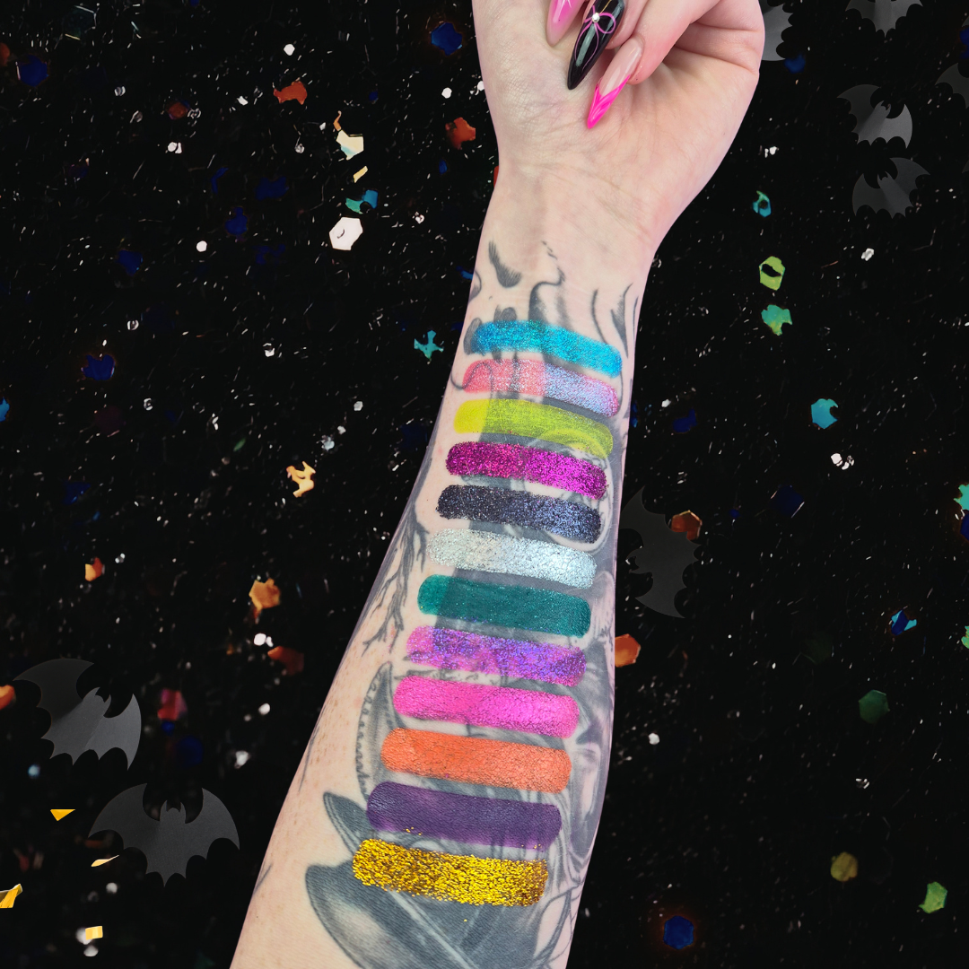 Colorful glittery makeup swatches on a tattooed arm with a black background and confetti.