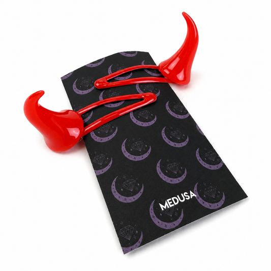 A pair of bright red horn-shaped hair clips with a glossy finish, displayed on top of their black branded packaging card featuring purple crescent moon and diamond patterns.