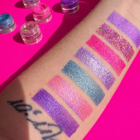 Glittery makeup swatches on a person's arm with open jars of glitter on a pink background
