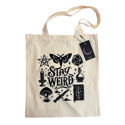 A cream-colored canvas tote bag with a black graphic print featuring occult symbols, a moth, a dagger, a potion bottle, a tarot card, a candle, and the words 'Stay Weird'. A black hangtag with a purple crescent moon logo is attached to the side.