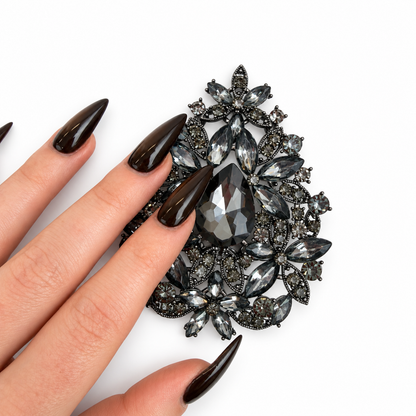 A close-up of a hand with long, glossy black manicured nails holding a large black teardrop-shaped rhinestone brooch against a plain white background.