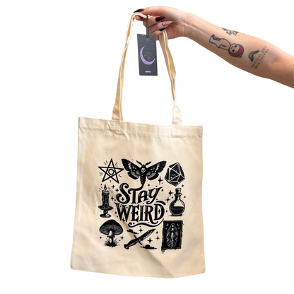 A cream-colored canvas tote bag with a black occult-themed graphic that reads 'Stay Weird' surrounded by illustrations like a moth, dagger, potion bottle, and pentacle. A hand with tattoos holds the bag by its handles against a solid white background.